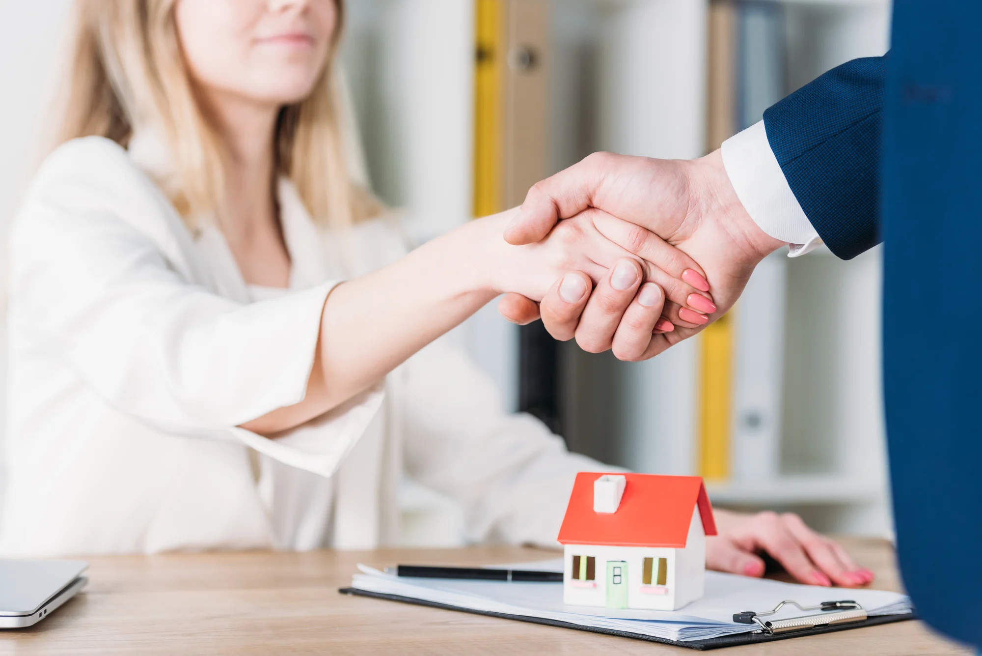 Partial view of smiling woman shaking hands with realtor near house model on table